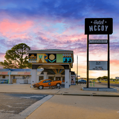 Un motel de carretera de estilo retro con un toldo pintado y un coche clásico naranja aparcado en la entrada, con un cielo de vivos tonos rosa y morado de fondo.