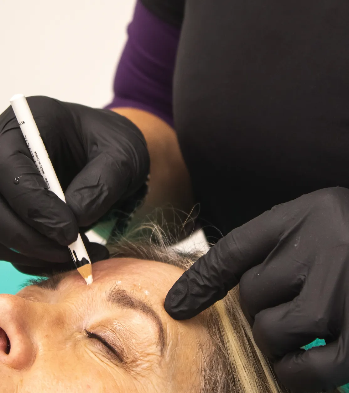 A practitioner in black gloves marking a client's forehead with a white pencil before treatment
