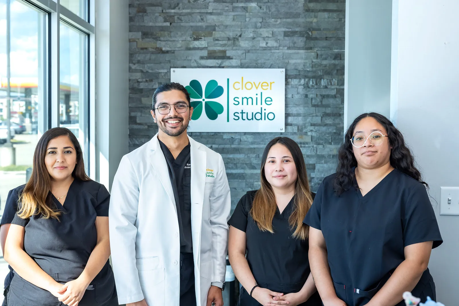 Dr. Karnik Shah, wearing a white dental coat, stands smiling with three female team members dressed in black scrubs at Clover Smile Studio. They are positioned in front of a stone accent wall featuring the Clover Smile Studio logo