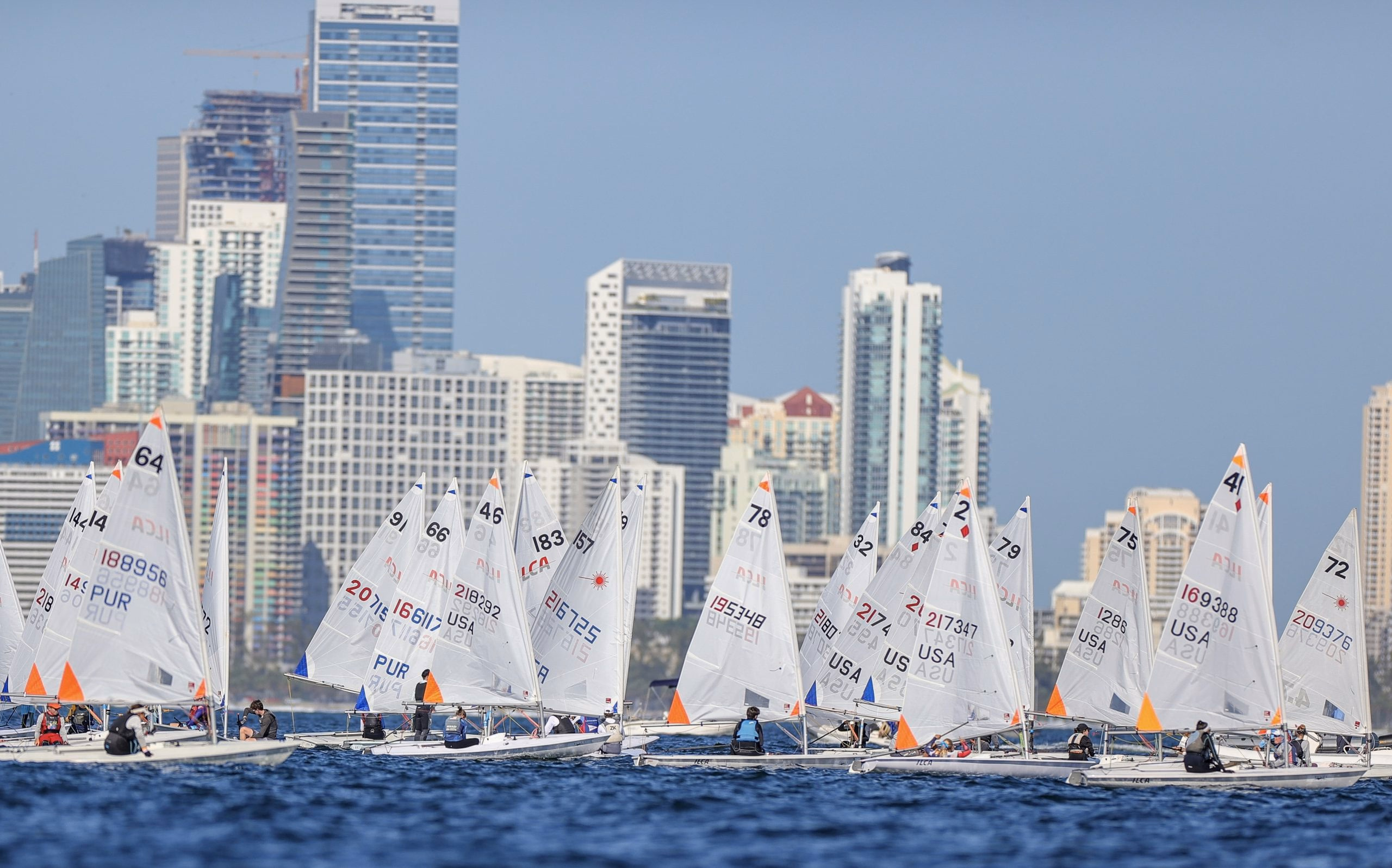 A fleet of one-design dinghies lined up on the water with the Miami skyline behind them
