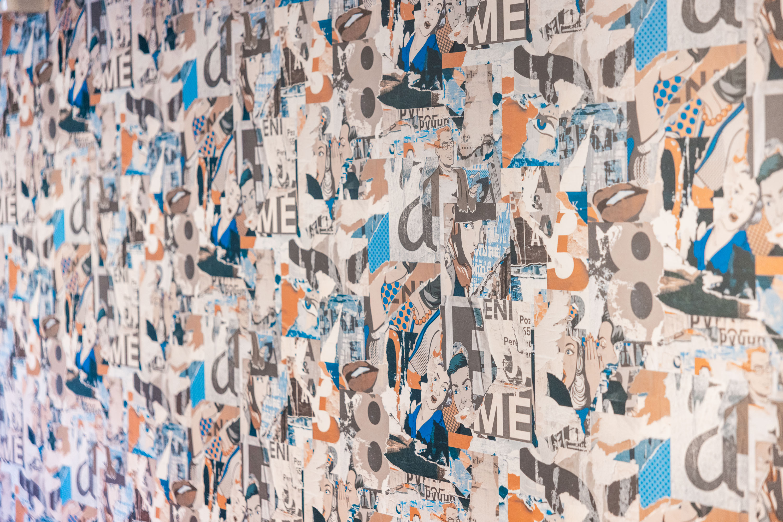 Wall covered with layered, torn posters featuring vintage comic-style faces, numbers, and letters in blue, orange, and beige tones.