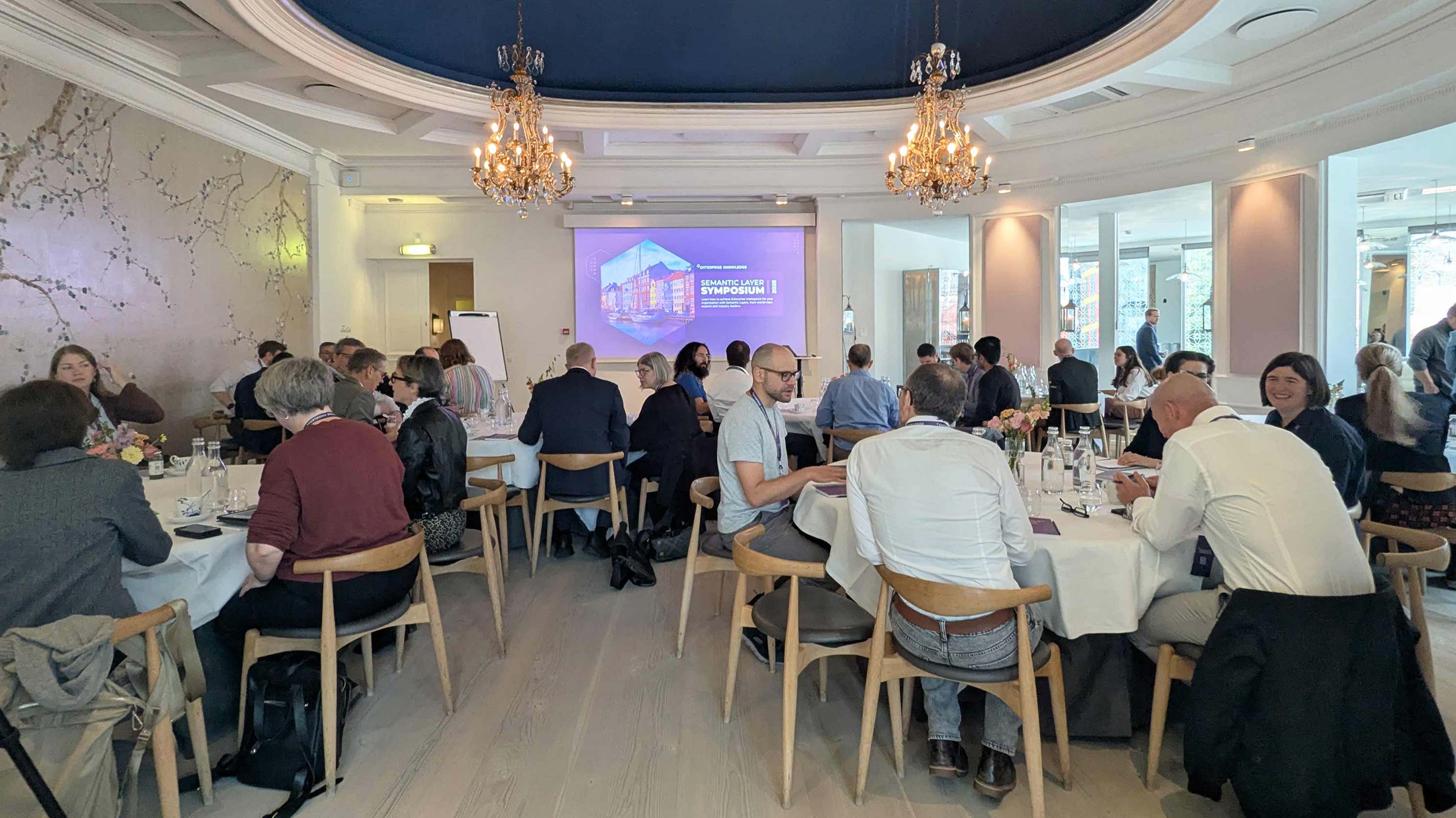 People seated around tables in a chandelier-lit room attending the Semantic Layer Symposium event.