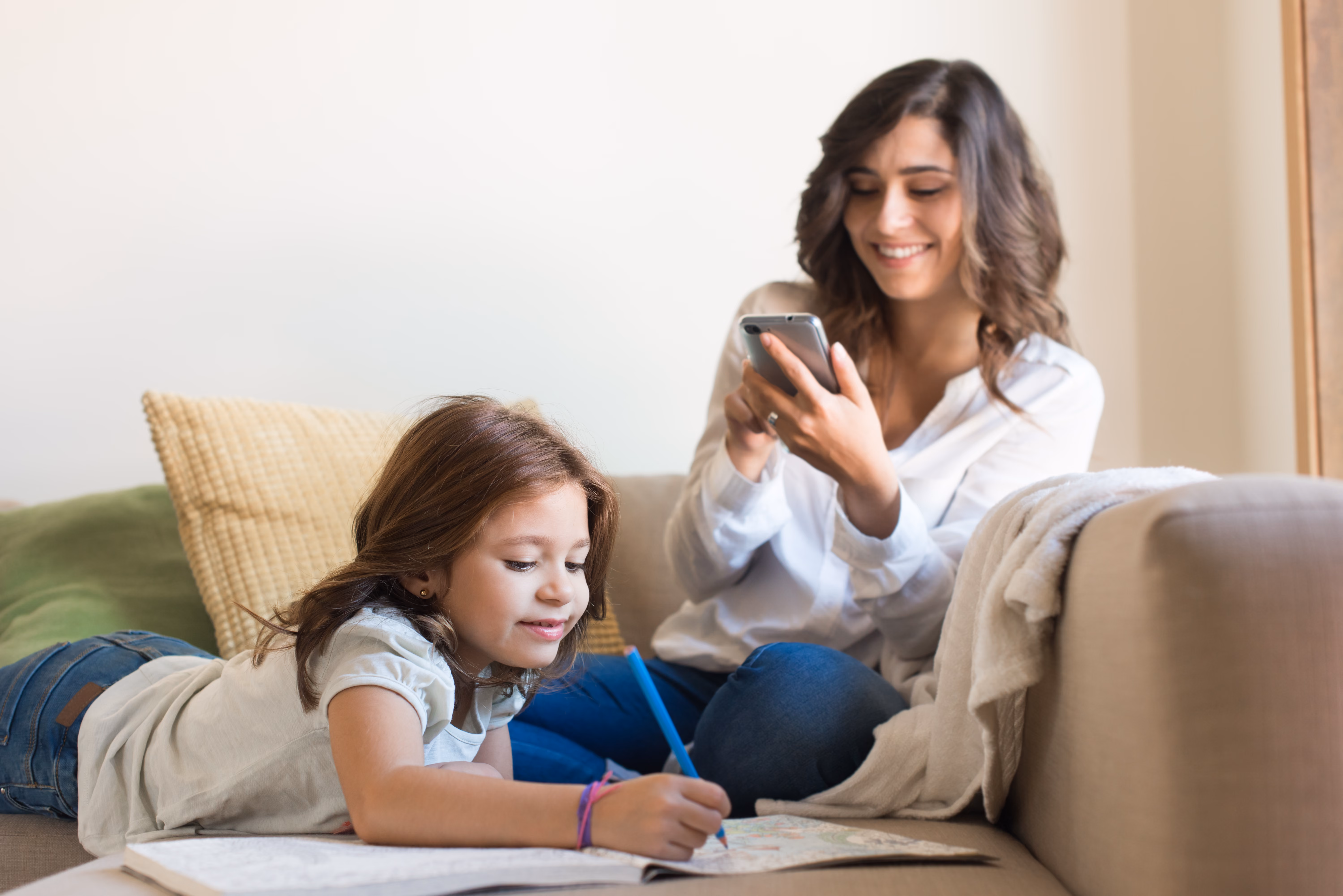 Young girl coloring in a book on a couch while a woman smiles looking at her phone beside her.