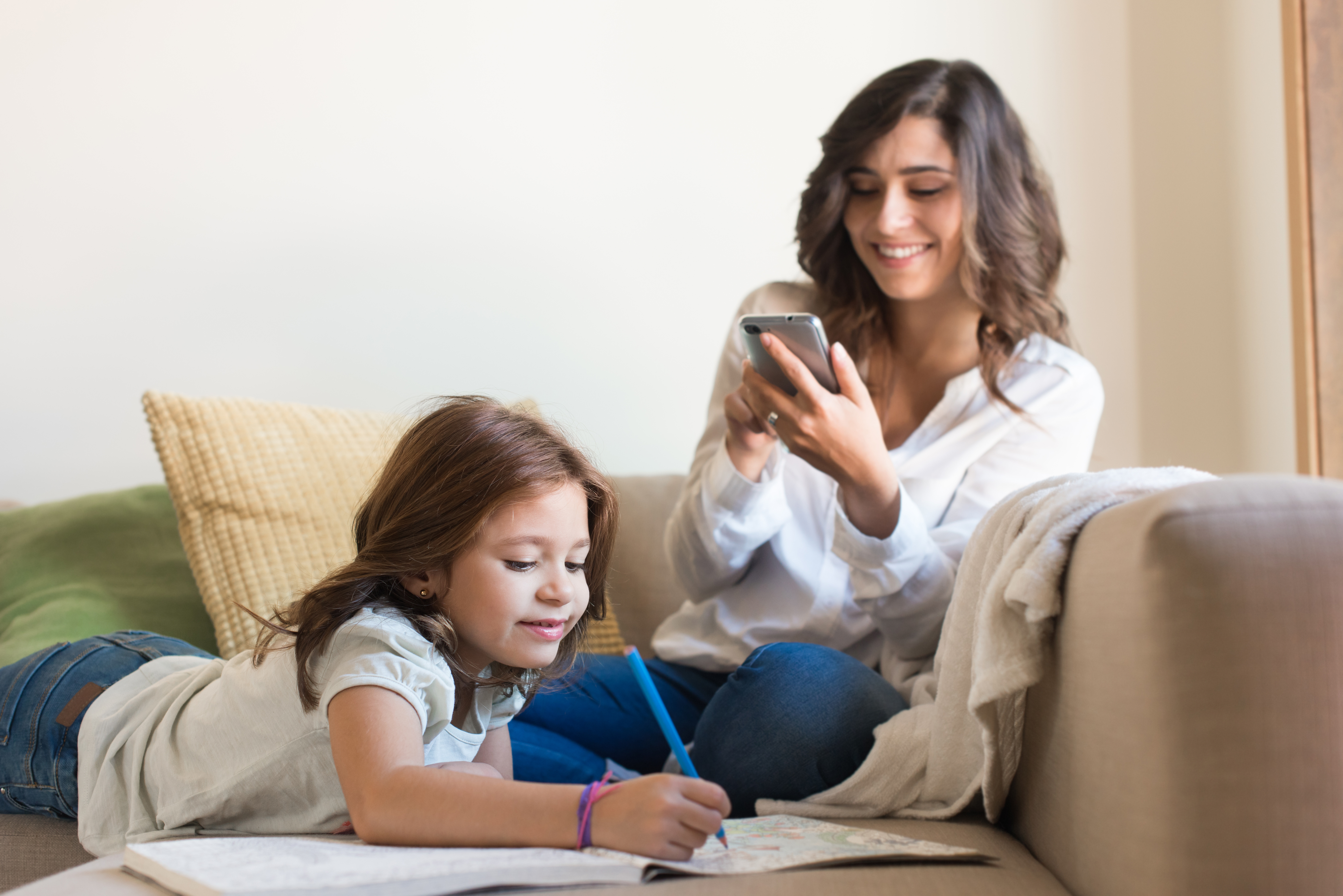 Young girl coloring in a book on a couch while a woman smiles looking at her phone beside her.