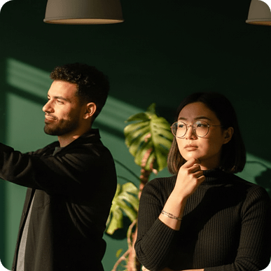 A man and woman stand in a room with green walls and sunlight, the man looking to the side and the woman wearing glasses and holding her chin thoughtfully.