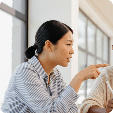 Woman with dark hair in a striped shirt pointing and speaking to another person indoors by a window.