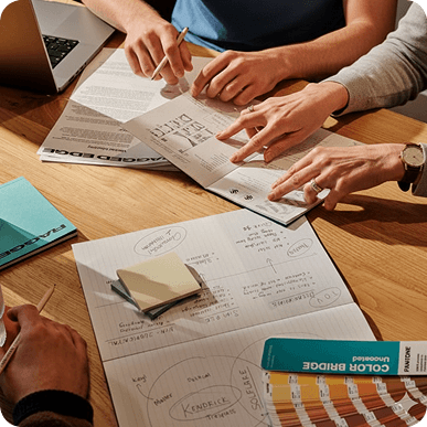Three people collaborating over a wooden table with documents, color swatches, a laptop, and notes.