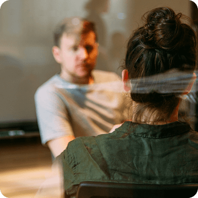 A man and a woman facing each other having a conversation, the woman is seen from behind and the man is slightly out of focus.