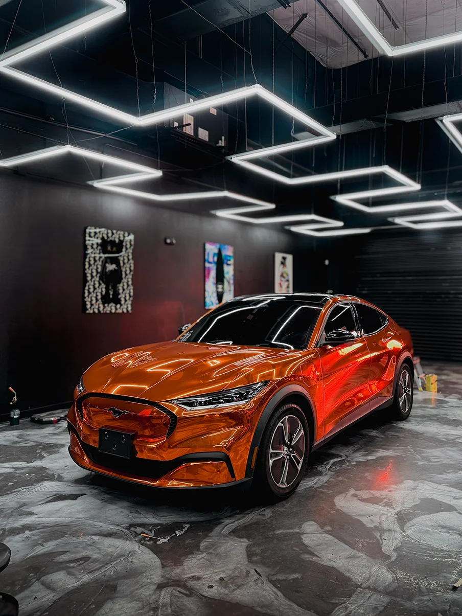 Vibrant gloss orange metallic car wrap on a Ford Mustang Mach-E, reflecting shop lights at Allure Auto Spa