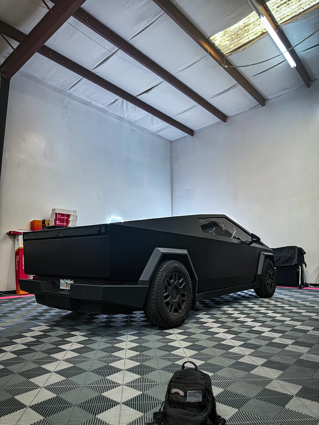 Rear three-quarter view of a matte black Tesla Cybertruck inside the Allure Auto Spa facility, showcasing its unique wrapped finish