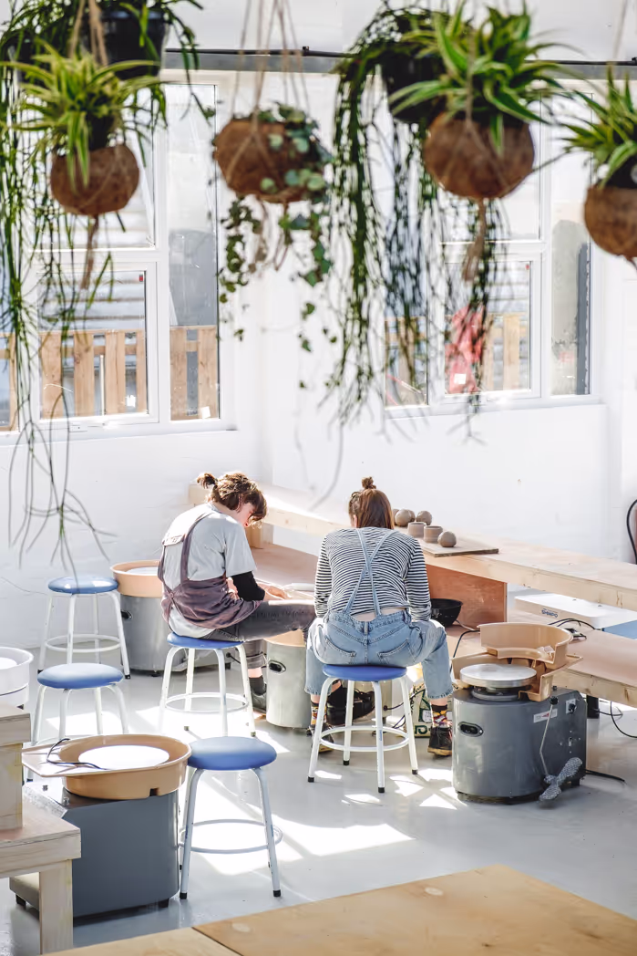 Two individuals seated at potter's wheels, surrounded by hanging plants from the ceiling, creating a serene atmosphere.