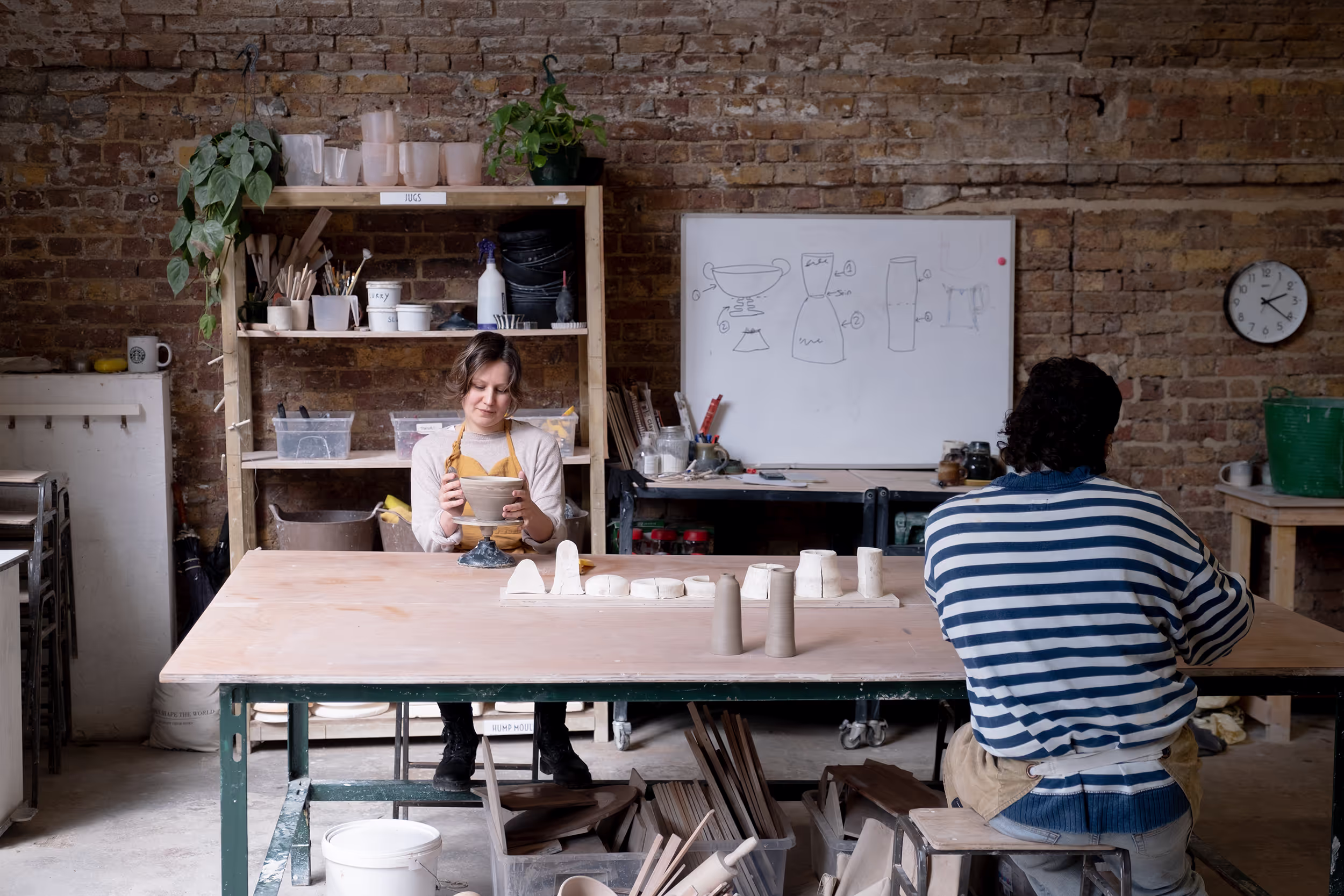 Two people working with clay in a pottery studio with tools and sketches on a whiteboard.