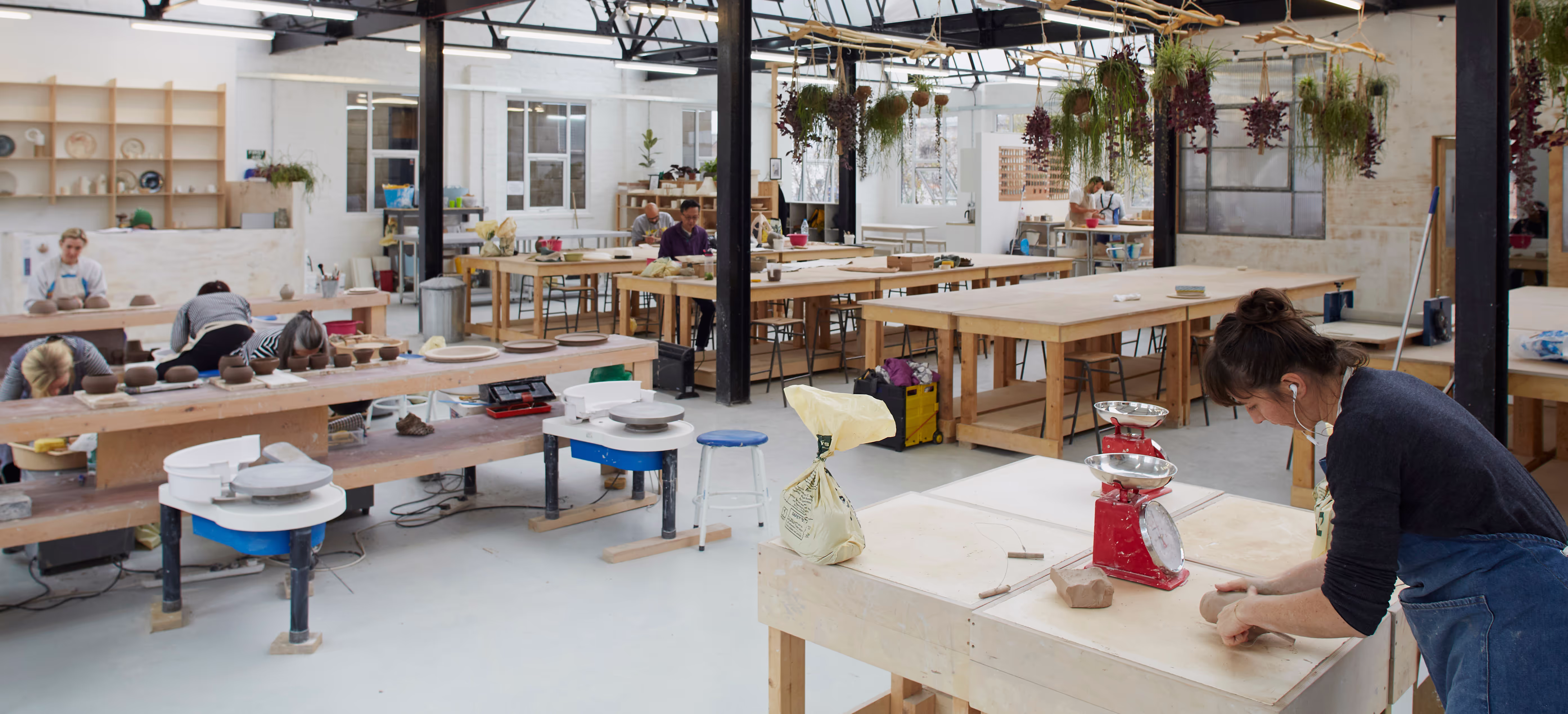 Pottery studio with several people working on clay projects at wooden tables under hanging plants.
