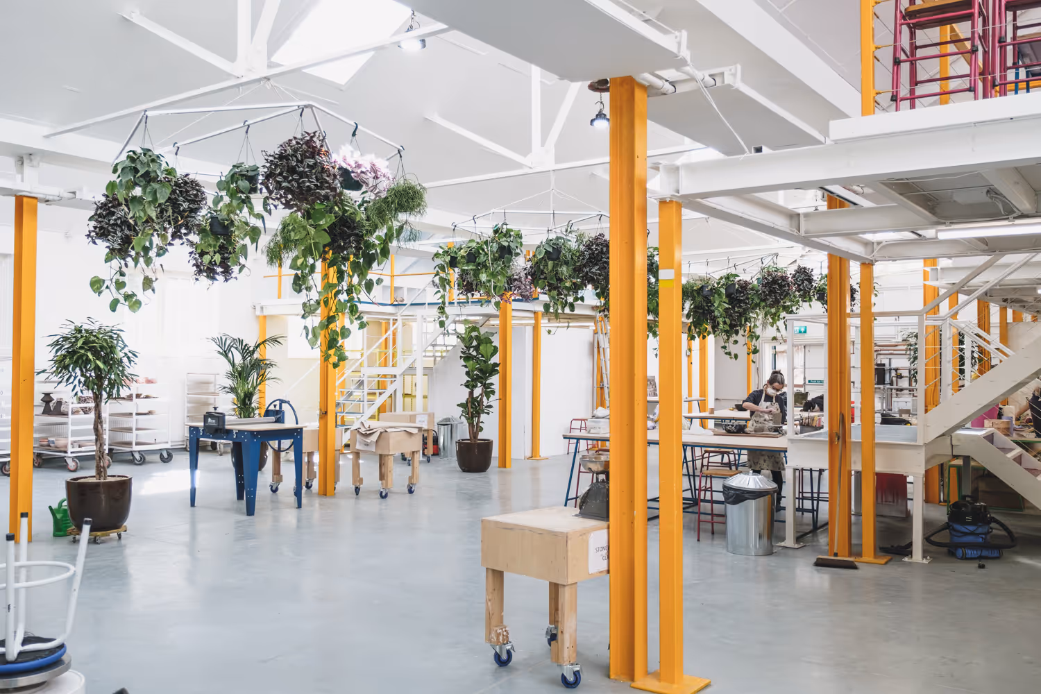 Bright spacious workshop with hanging plants, yellow support beams, work tables, and a person working.