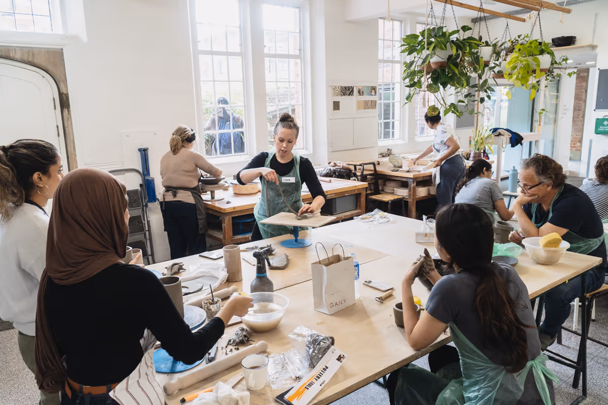 Group of people crafting pottery in a bright room with large windows and hanging plants.