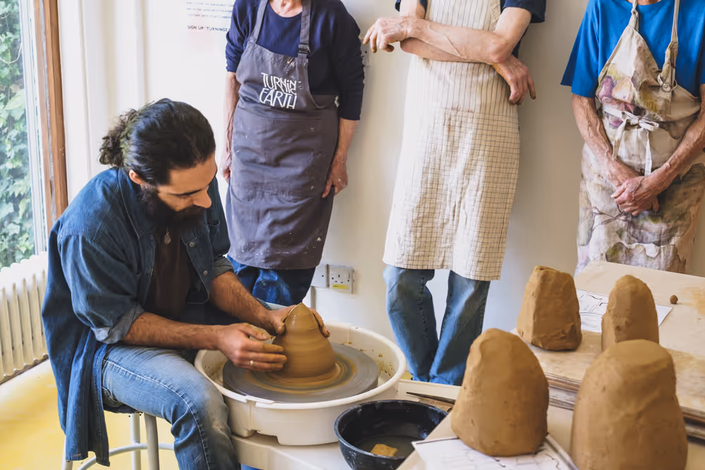 Man shaping clay on potter's wheel while three people observe in aprons in a pottery studio.