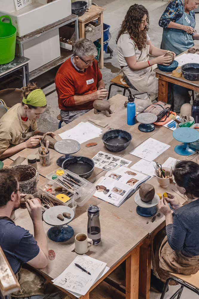 Students working on pottery around a table in a well-lit studio with large windows