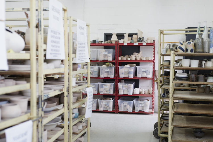 Shelves of members' pottery at Leyton: wooden racks of unglazed work and red shelving with labelled storage boxes