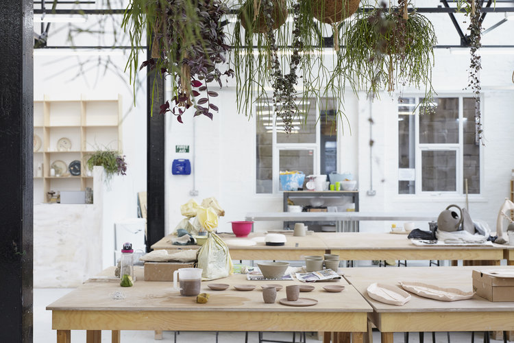 Hand-building tables at Leyton laid out with work in progress, tools and clay, under trailing plants