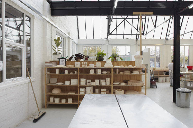 Shelves of unglazed pots and bowls beside a steel worktable, under the glass roof of the Leyton studio