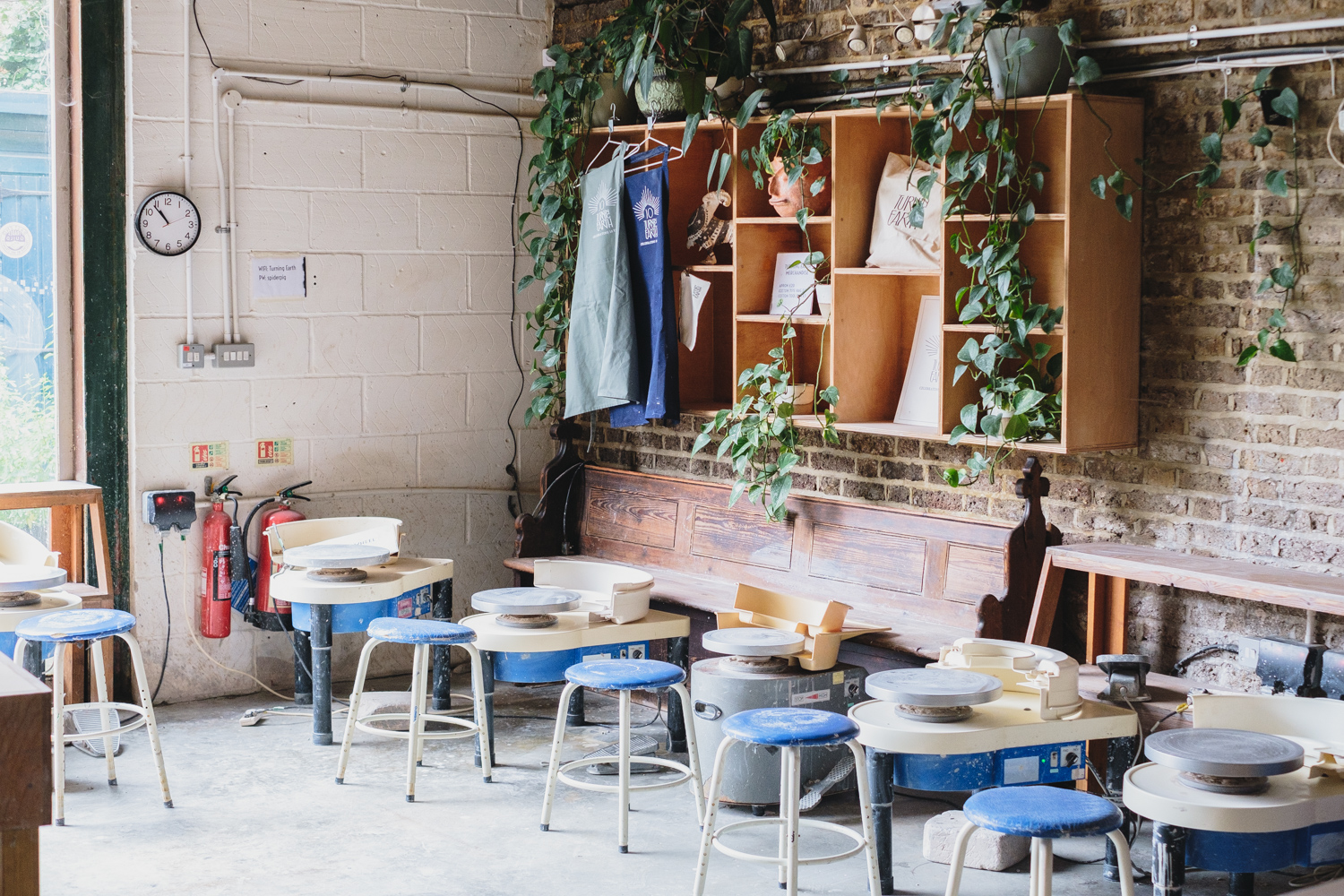 A row of pottery wheels with blue stools at Hoxton, beside an old wooden pew and shelves trailing with plants