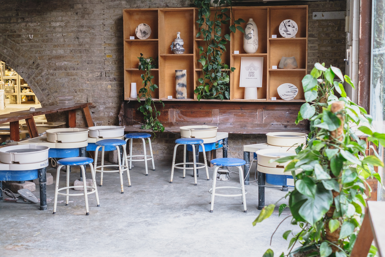 Pottery wheels and blue stools beneath wooden shelves of finished, decorated pots at the Hoxton studio
