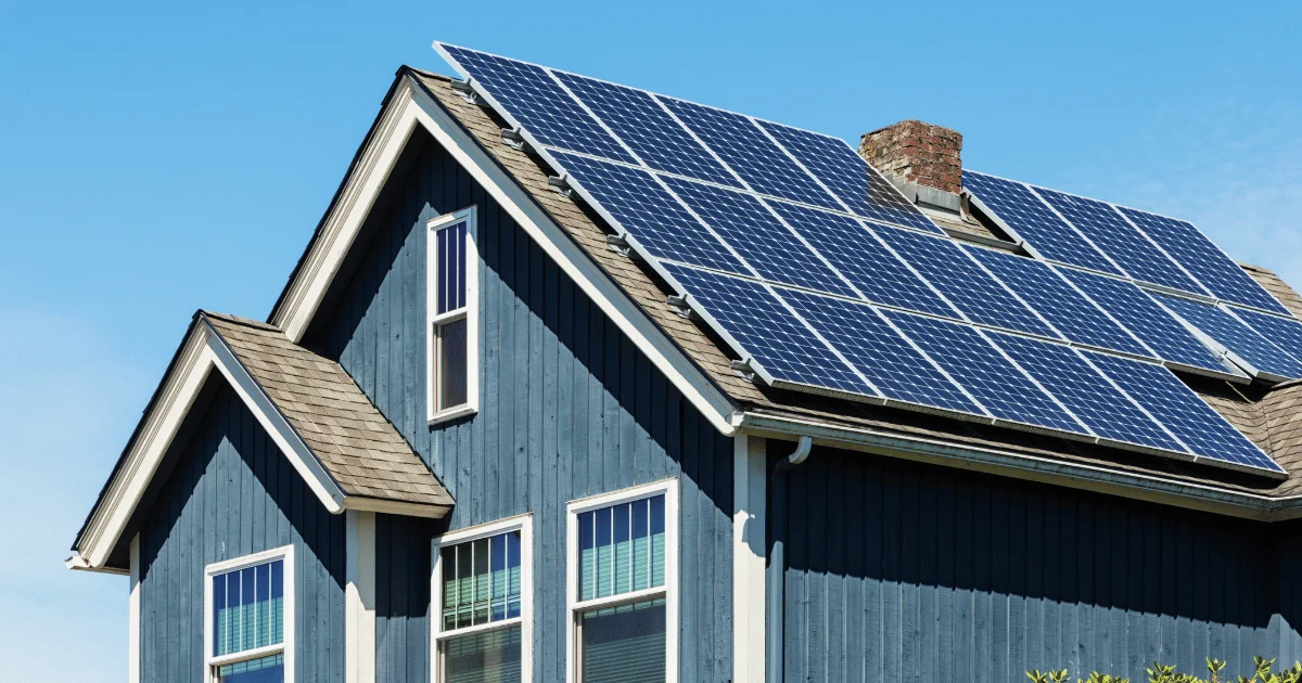A bright exterior photograph of a residential home with blue siding and a roof fully equipped with a small-scale solar photovoltaic array against a clear sky.