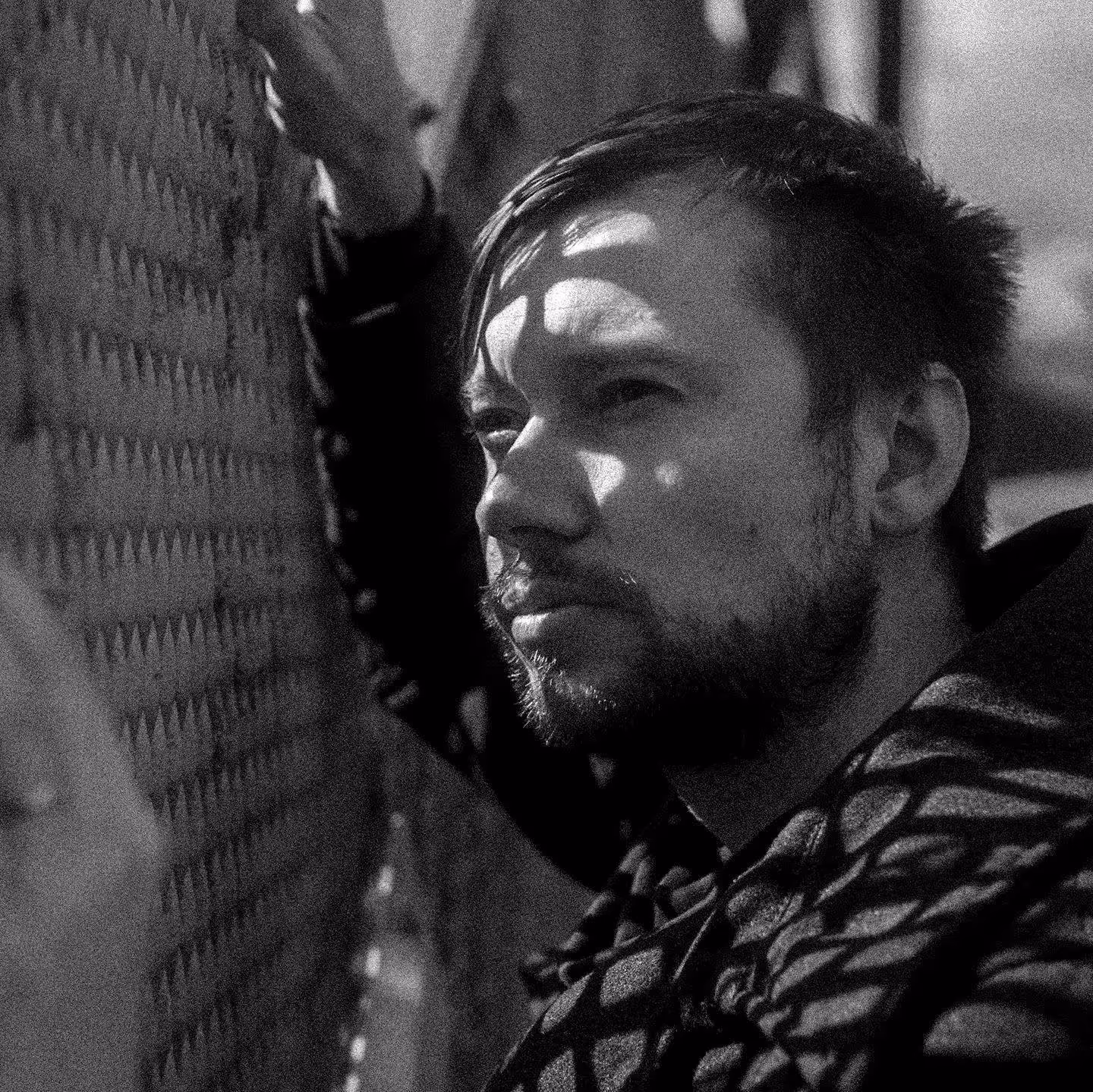 Black and white close-up portrait of a man with a beard resting his arm on a textured metal fence, with shadows cast on his face.