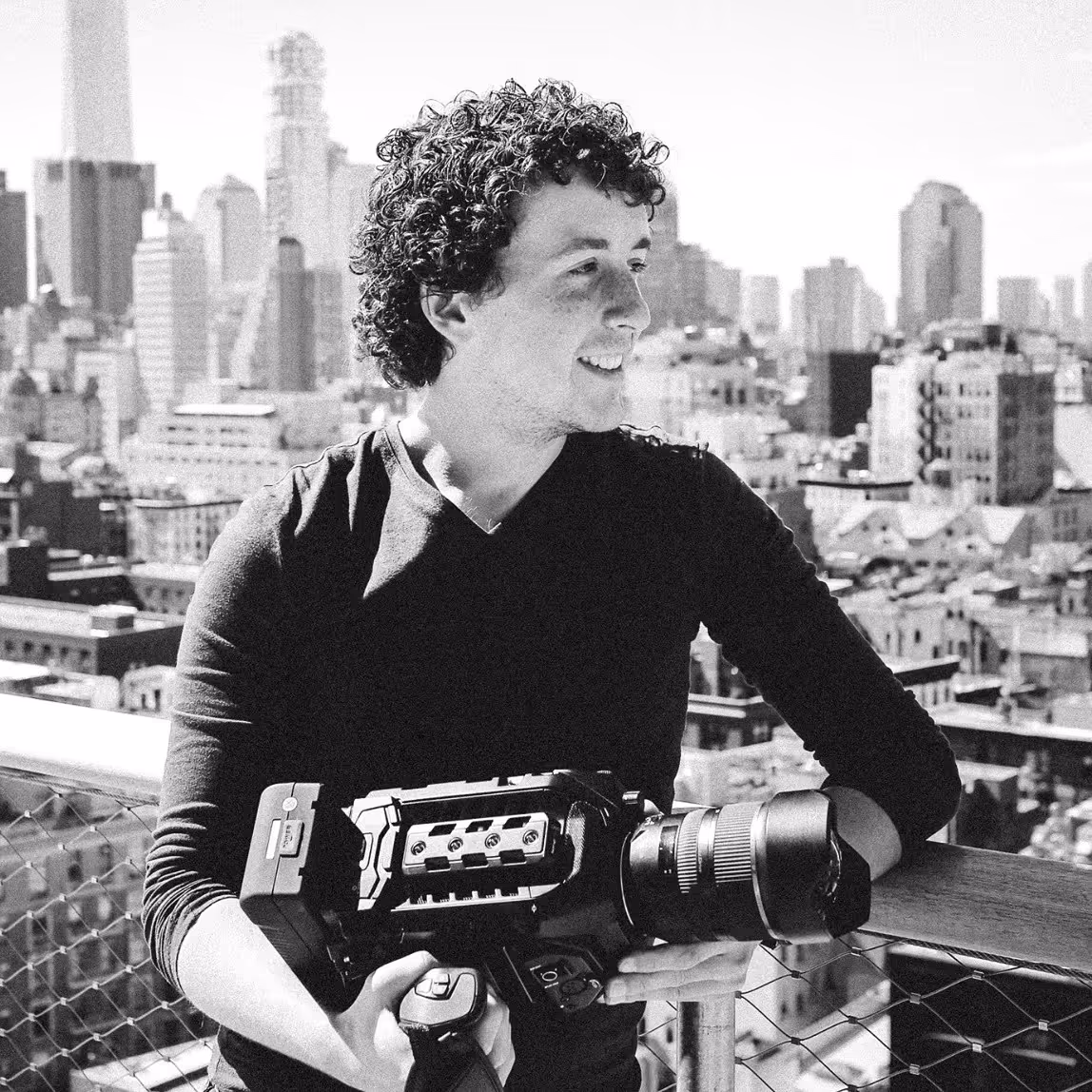 Smiling man with curly hair holding a professional video camera on an outdoor balcony overlooking a city skyline.