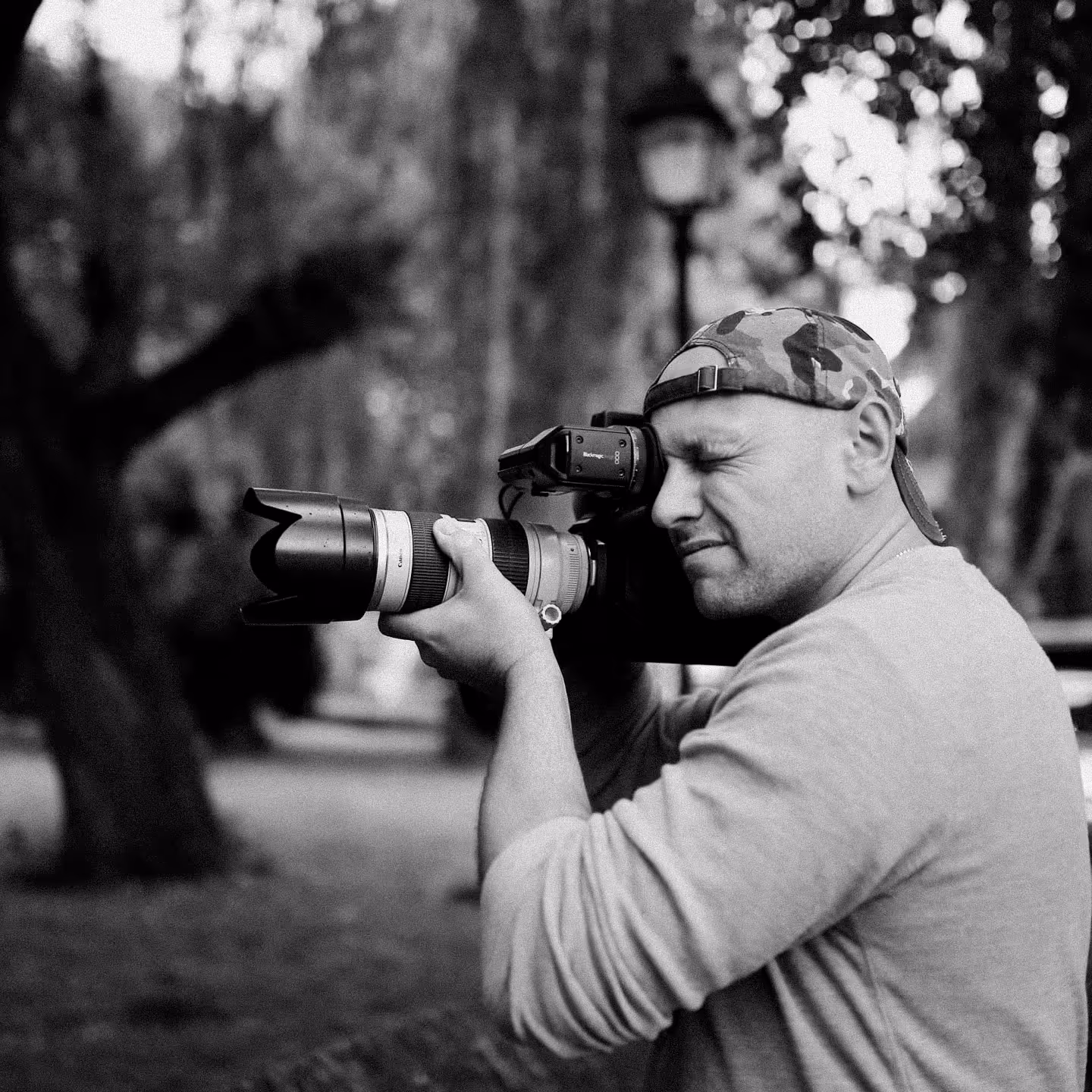 Man wearing a camouflage cap backward, using a professional camera with a large zoom lens in an outdoor setting.