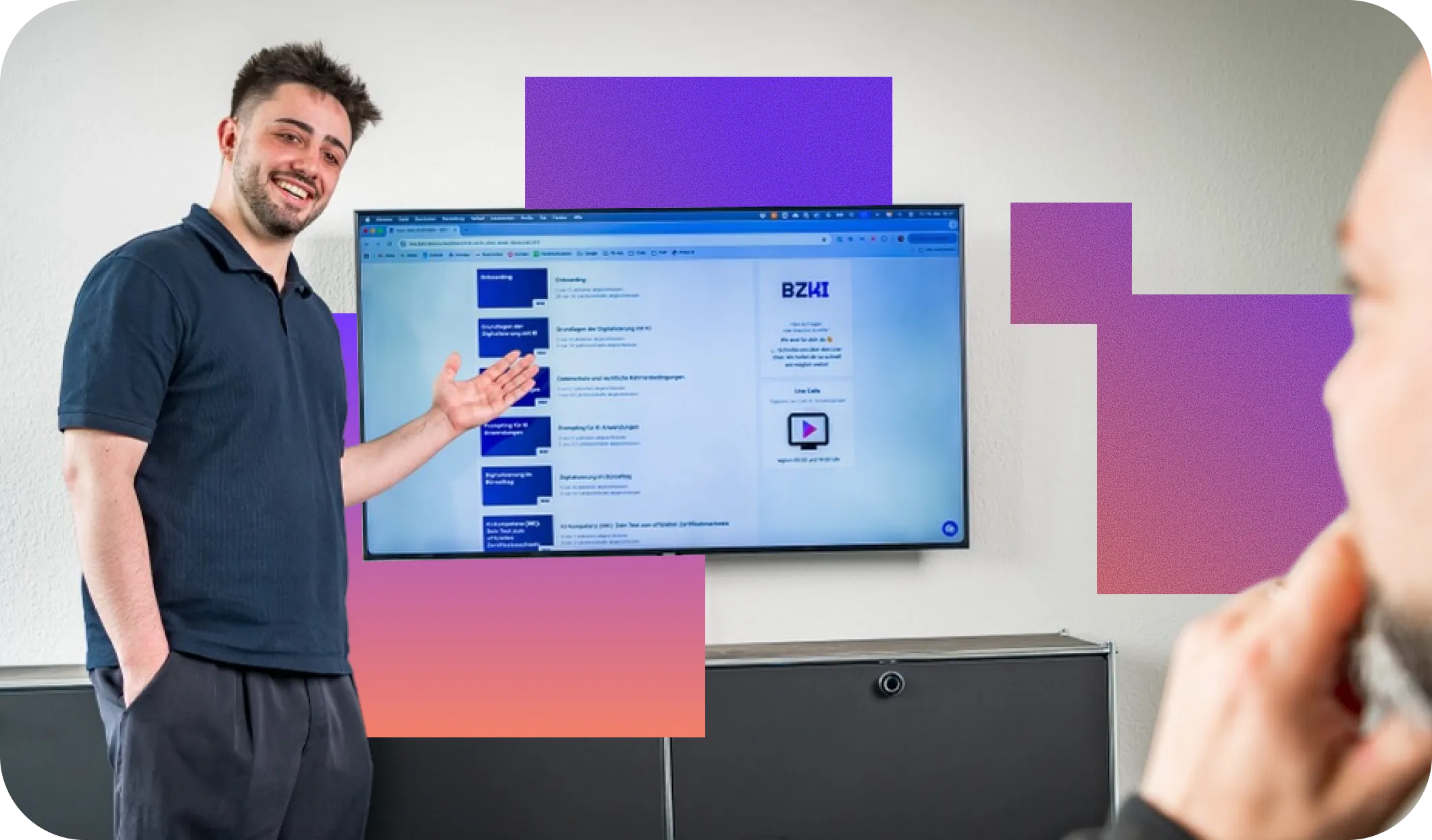 A man in a navy blue shirt smiling and presenting a screen on the wall displaying a webpage with blue navigation blocks and text, while another person with a beard listens attentively.