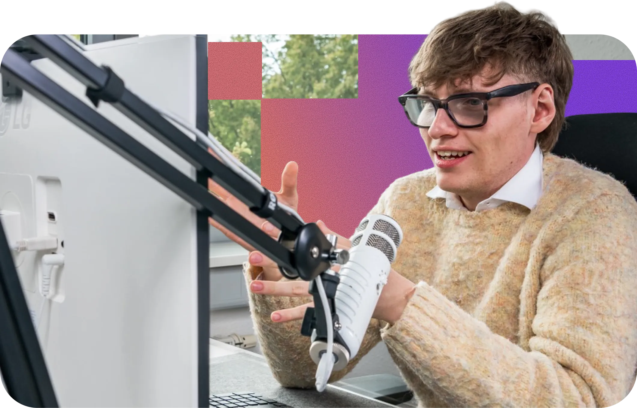 Young man wearing a beige sweater and large dark glasses speaking into a white microphone mounted on an adjustable arm in front of a computer monitor at a desk.