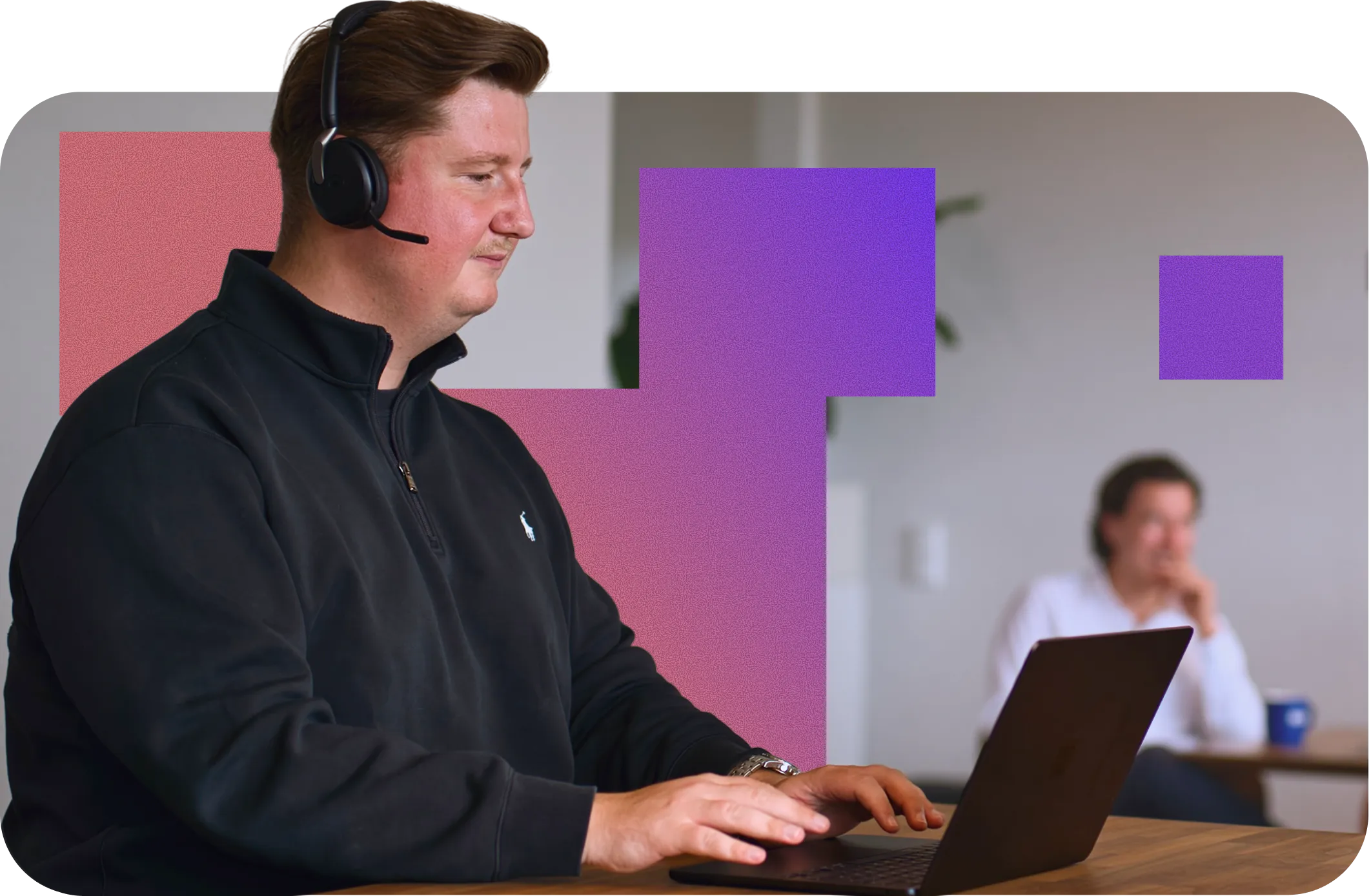 Man wearing a headset and black zip-up jacket typing on a laptop at a wooden table, with another person blurred in the background.