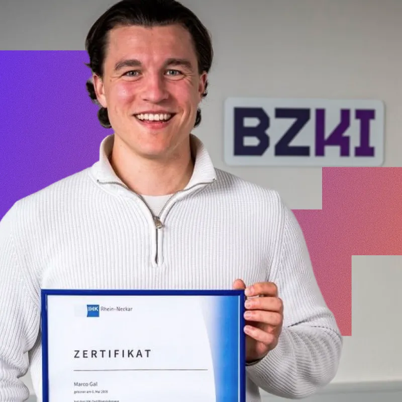 Smiling man with dark hair wearing a white sweater holding a certificate in a blue frame, standing indoors with a wall displaying the logo 'BZKI' in the background.