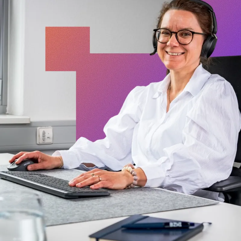 Smiling woman wearing glasses and headphones, sitting at a desk using a keyboard and mouse in a bright office.