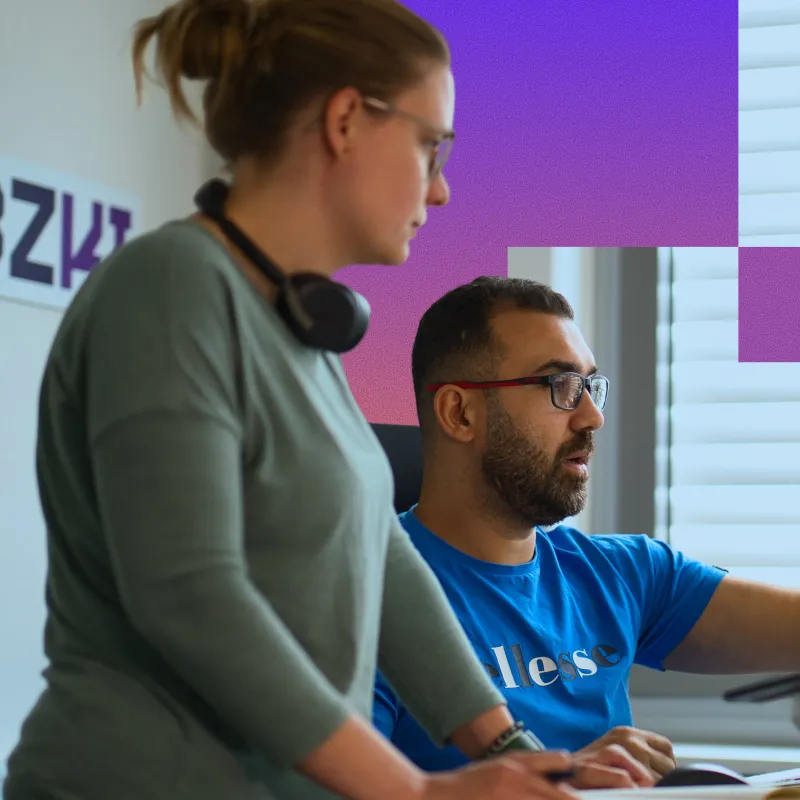 A man with glasses in a blue shirt and a woman with a bun and headphones around her neck are working together at a computer in an office setting.