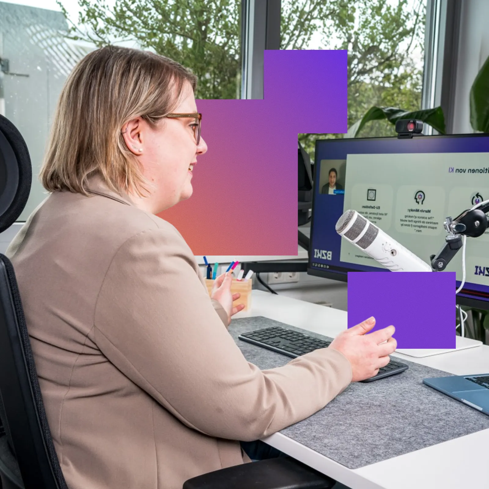 Woman with short hair and glasses sitting at a desk with a microphone and keyboard, speaking while looking at a computer screen.