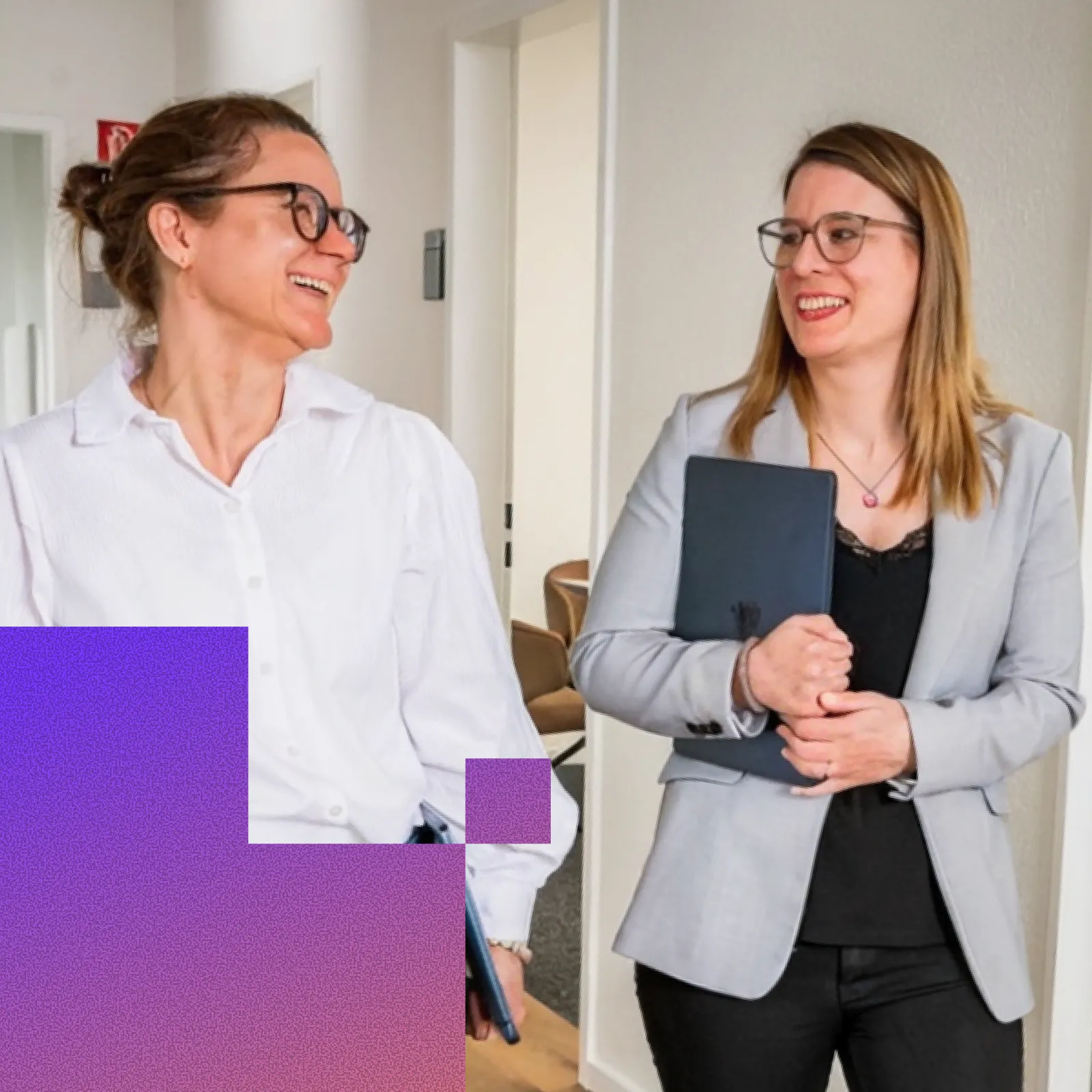 Two professional women wearing glasses smiling and talking in an office hallway, one holding a tablet and the other holding a folder.