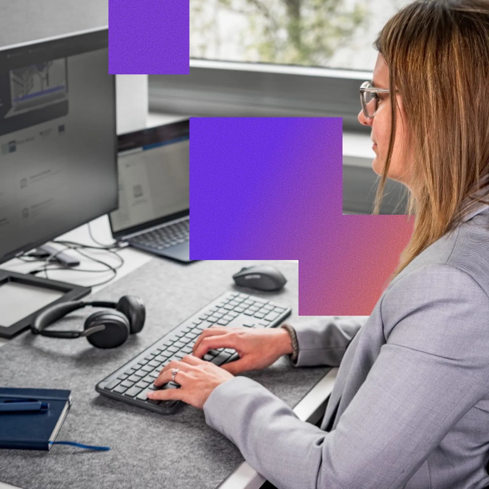 A woman wearing glasses and a gray blazer is typing on a keyboard at a desk with a laptop, a monitor, headphones, and a mouse, with a window in the background.