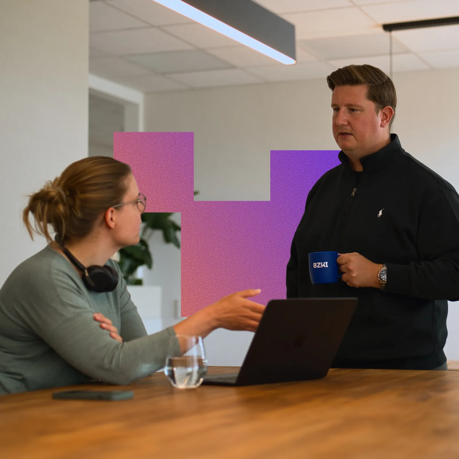 A man holding a blue BZKI mug stands and talks to a seated woman with glasses and headphones around her neck, who gestures toward a laptop on a wooden table.
