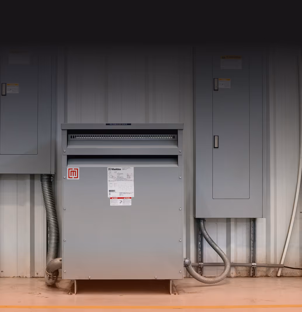 Industrial electrical panel with warning labels and gray metal conduits attached mounted on a wall in a utility room.