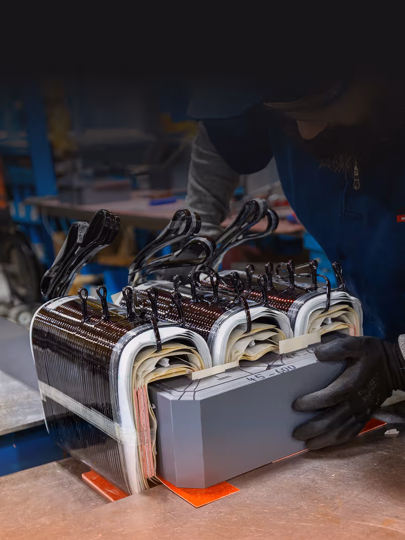 Worker assembling or inspecting large industrial transformer coils on a metal workbench.
