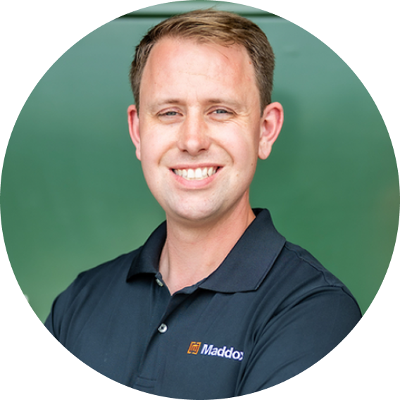 Smiling man with short light brown hair wearing a black Maddox polo shirt against a green background.