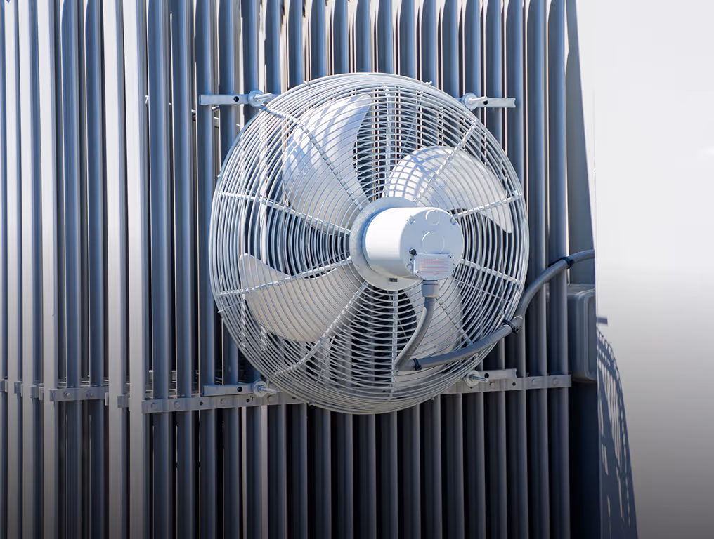Industrial metal ventilation fan with protective grille mounted on gray corrugated wall.