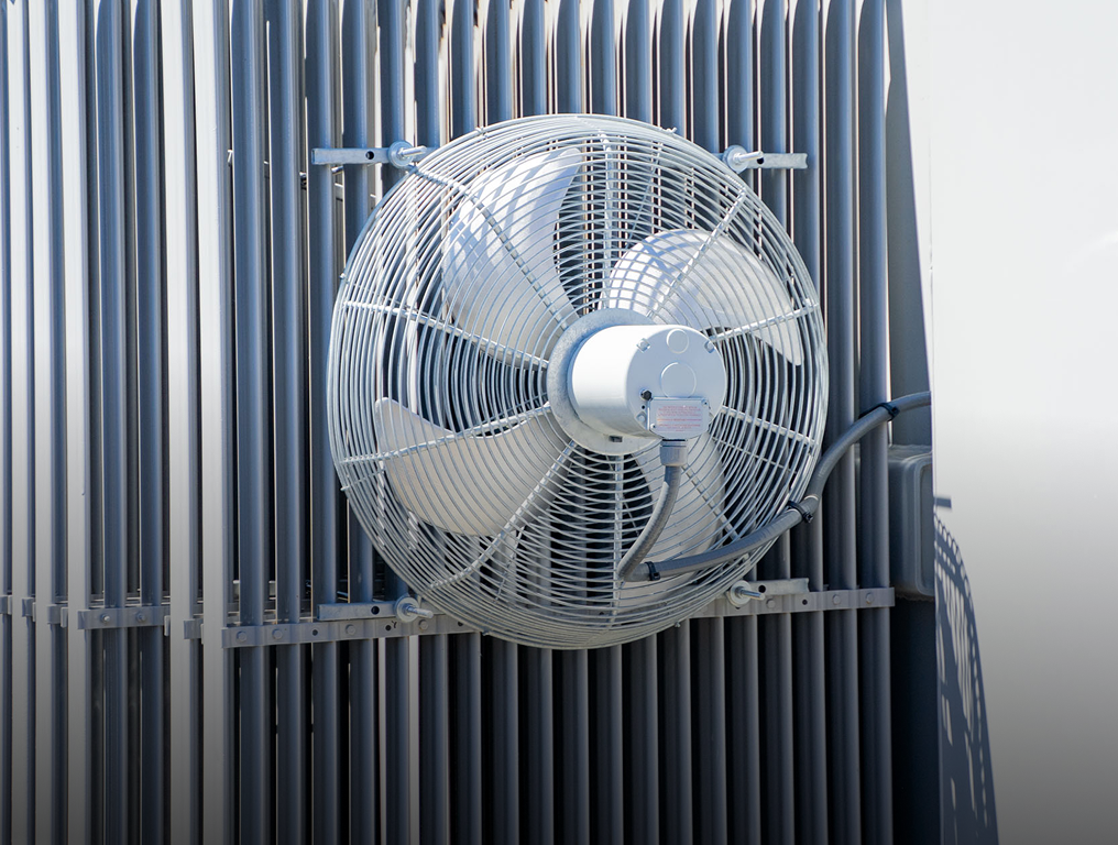 Industrial metal ventilation fan with protective grille mounted on gray corrugated wall.
