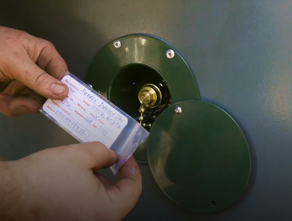 Hands holding a laminated tag next to a green metal circular electrical connector on a wall.
