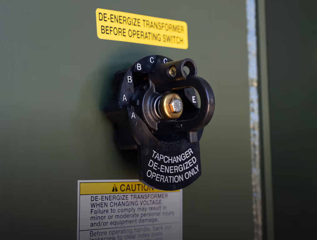Close-up of transformer tap changer with caution labels about de-energizing before operating.