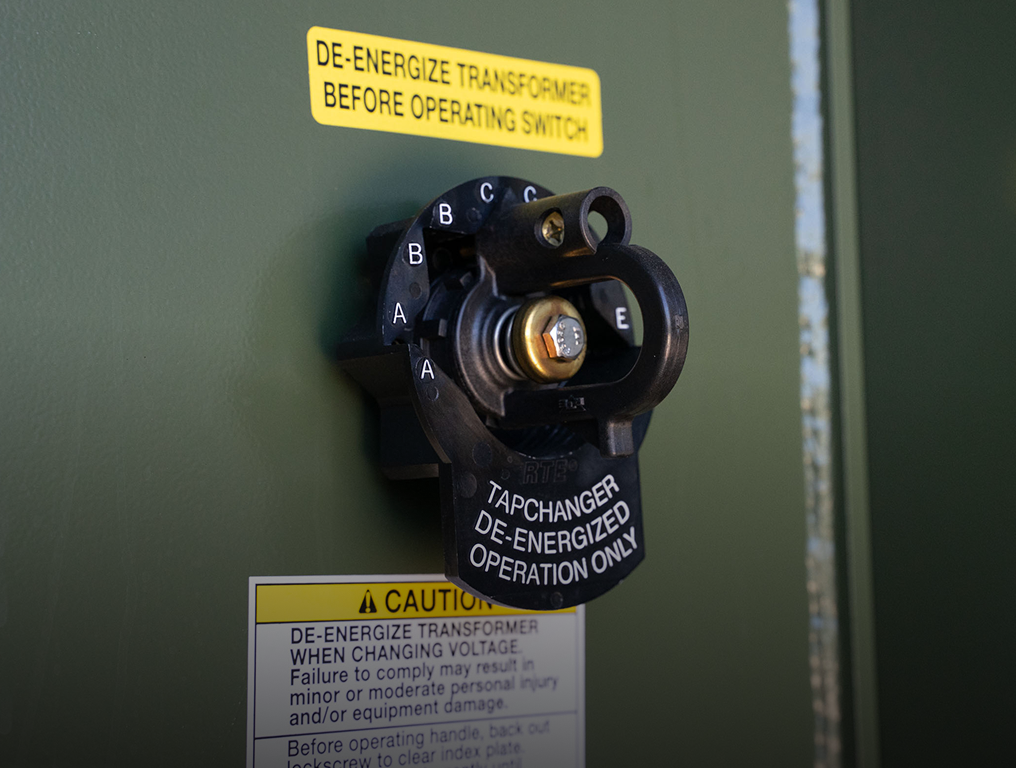 Close-up of transformer tap changer with caution labels about de-energizing before operating.