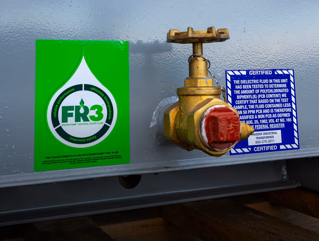 Close-up of a transformer valve with green FR3 fluid and blue certification labels on a gray metal surface.