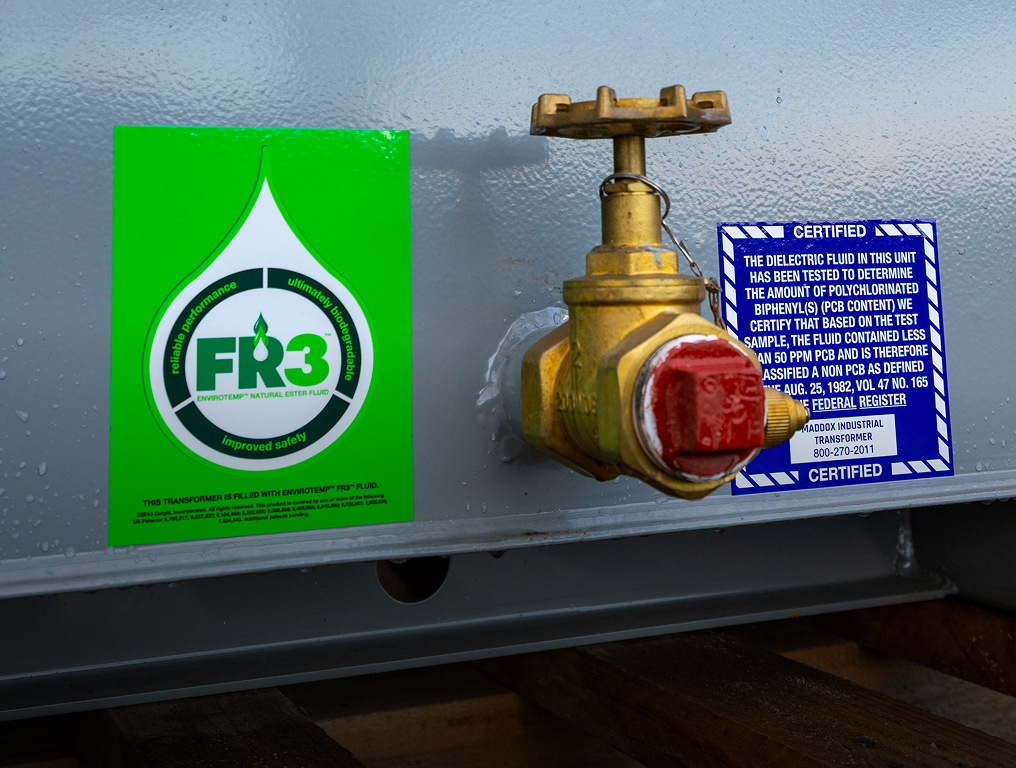 Close-up of a transformer valve with green FR3 fluid and blue certification labels on a gray metal surface.